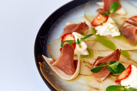A gourmet plate featuring thinly sliced prosciutto arranged elegantly with fresh strawberries, creamy cheese, and a drizzle of vibrant sauce on a modern dish.の写真素材