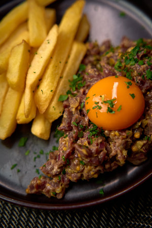 A delectable dish featuring a rich meat tartare topped with a vibrant egg yolk, accompanied by crispy golden fries and fresh herbs on a stylish plate.の写真素材