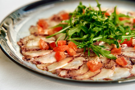 A Deliciously Prepared Plate of Thinly Sliced Octopus Carpaccio Garnished with Fresh Tomatoes and Arugula on a Stylish Plateの写真素材