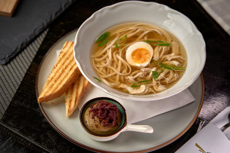 A Delicious Bowl of Clear Broth Noodle Soup with Tender Chicken, Soft Boiled Egg, Fresh Herbs, and Crispy Toasted Bread on a Modern Plateの写真素材