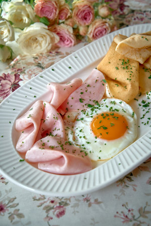 Delicious Plate of Ham Slices, a Fried Egg, and Crispy Pancakes Garnished with Fresh Herbs on a Floral Table Settingの写真素材