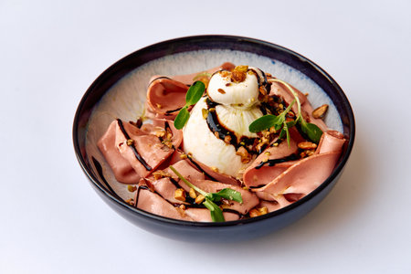 Deliciously Arranged Plate of Creamy Burrata Cheese Served with Delicate Slices of Meat Garnished with Nuts and Fresh Greens in a Stylish Dishの写真素材