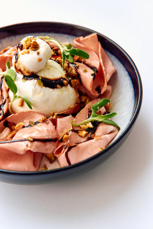 An Artistic Culinary Creation Featuring Creamy Burrata Cheese Surrounded by Thinly Sliced Ham, Garnished with Nuts and a Drizzle of Balsamic Reductionの写真素材