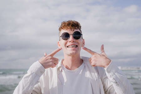 a young guy shows his fingers on the braces on his teeth, with curls on his head and sunglassesの写真素材