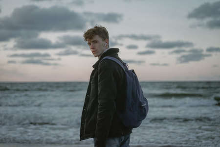 a young guy with a backpack behind his back stands against the backdrop of a stormy sea, looks into the camera, curly hair on his head, in a warm jacket, cold seasonの写真素材