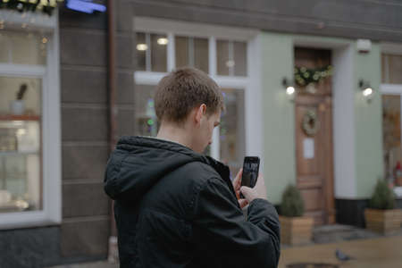 a guy in a jacket photographs a city street on a mobile phone, holds a smartphone in his hands, modern generationの写真素材