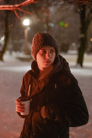 a young man in a gloomy winter forest, in a knitted warm hat, snow around, holding a warming drink...