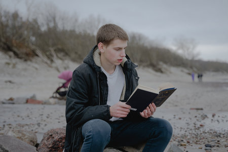 teenager sits on the beach reads a book, looks into a textbook. immersed in the study of the materialの写真素材