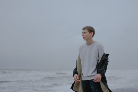 a teenager in a white sweatshirt stands against the background of the cloudy sea, looks thoughtfully...