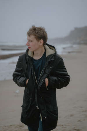 a guy in a winter jacket walks along the beach, grinds to the side, a teenager travels, a strong wind blows in his face from the seaの写真素材