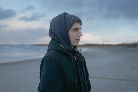 portrait of a pensive teenager, a young man in a hat on the seashore, a guy's journey in the winterの写真素材