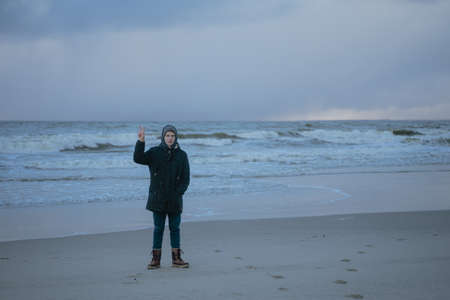 cold stormy winter sea, a small man stands in full growth with his hand raised with two fingersの写真素材