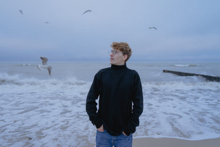 seagulls are flying, a young man in a sulfur walk stands on the shore of a stormy sea with an...