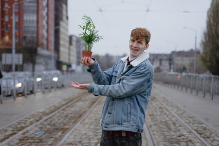 a man in a blue denim jacket holds a pot with a green flower in his hand and laughs mischievously, a smile with braces, with curly hair on his head, stands on the streets of passing cars with exhaust gases, an ecological problem of the cityの写真素材