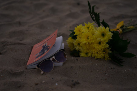 a book with sunglasses and a bouquet of yellow flowers lies on a sandy beach, important accessories during a holiday at seaの写真素材