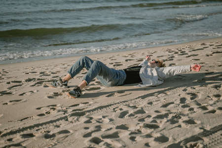 the guy fell on the sandy beach enjoys a sunny day by the sea, laughs while lying on the sandの写真素材
