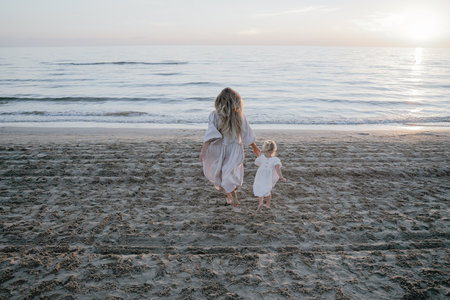 mother and daughter running along the sandy beach in white dressesの写真素材