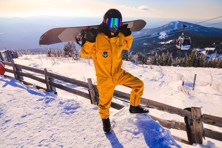 a snowboarder in a yellow overall stands on top of a mountain with a board thrown back over his shoulders. Sheregesh city 09.02.2023の写真素材