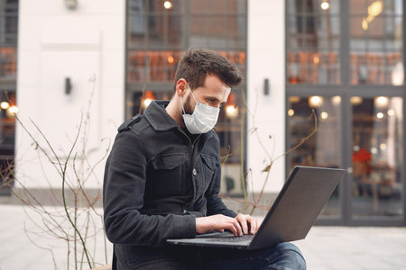 Man in a mask sitting in the city with a laptop