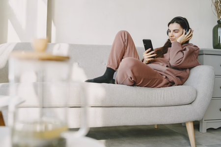 Woman at home with headphones and phone communicates onlineの写真素材