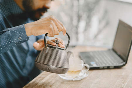 Man sitting in a cafe and working behind a laptop, writing in a notebook and pouring a tea in a cup.の写真素材