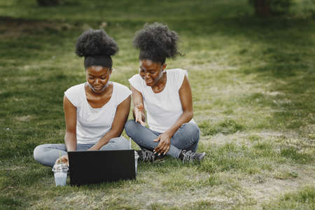 Young african-american twin sisters sitting on a grass in a park and using a laptopの写真素材