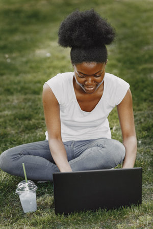 Portrait of a smiling young african-american girl sitting on a grass with a laptopの写真素材