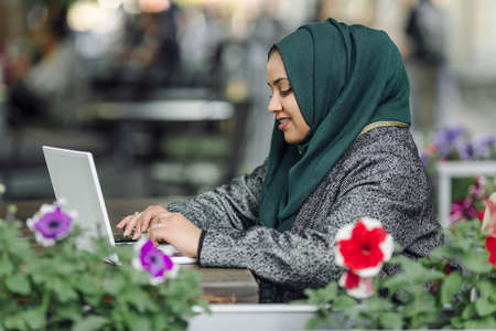 Portrait of a muslim girl in a green hijab with a laptop in the street cafeの写真素材