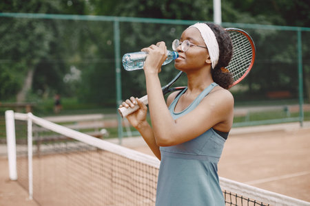 Black american female tennis player drinking water on the court outdoorsの写真素材