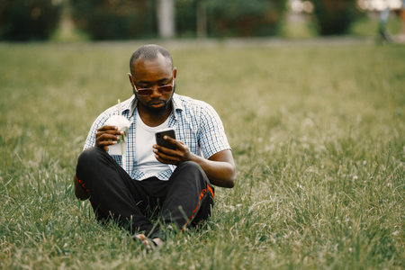 Black american man in a sunglasses sitting on a grass and using a phone