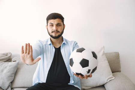 Brunette guy sitting on a coach with a ball and showing sign stopの写真素材