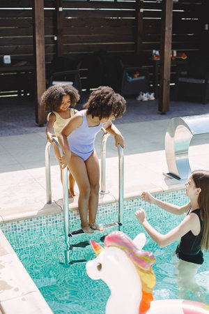 Two african american sisters standing near pool stairs and mother helping them