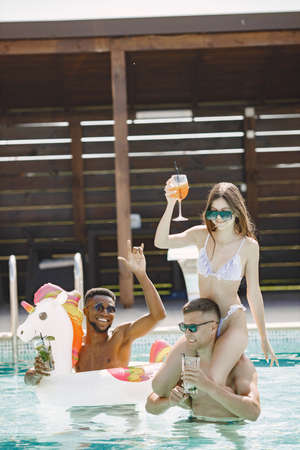 Three friends in a swimwears relaxing in the swimming pool and drinking a cocktailsの写真素材