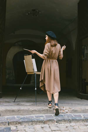 Girl in dress and black hat painting on an easel in a city on a streetの写真素材