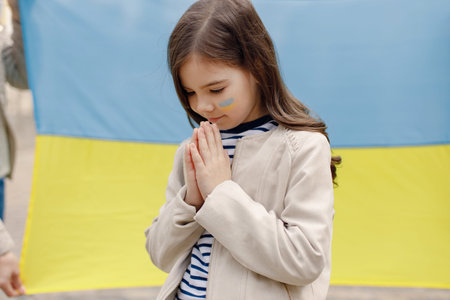 Little girl with a flag of Ukraine on a cheek praying in front of big flag