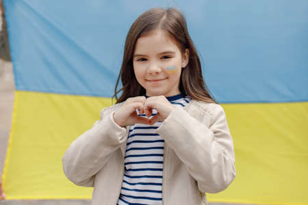 Little girl with a flag of Ukraine on a cheek show a heart in front of big flagの写真素材