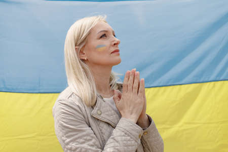 Woman with a flag of Ukraine on a cheek praying in front of big flagの写真素材