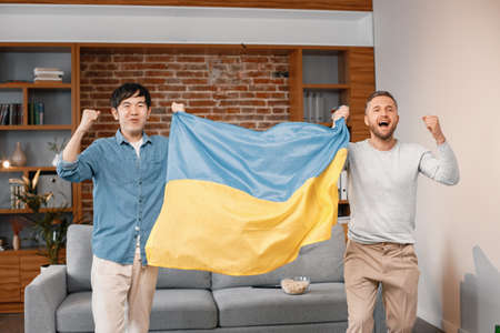 Two men watching a football game on tv and holding big flag of Ukraineの写真素材