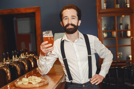 Man with a mustache and beard stands at the bar and drinks alcohol from a glass.の写真素材