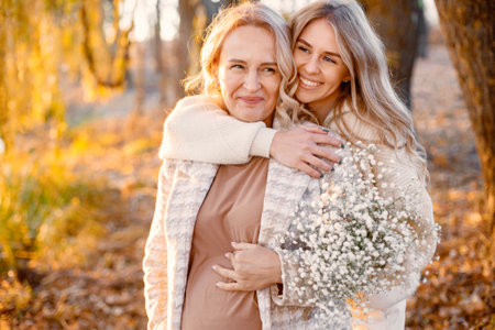 Adult daughter hugs her pregnant mom while standing in autumn parkの写真素材