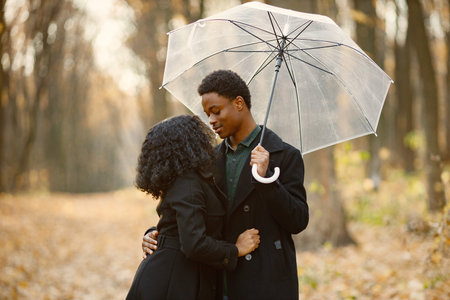 Loving black couple walking in park on autumn day holding a transparent umbrellaの写真素材