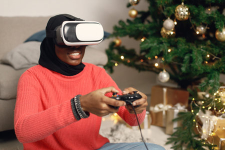 Muslim girl in black hijab sitting near Christmas tree wearing virtual reality glasses