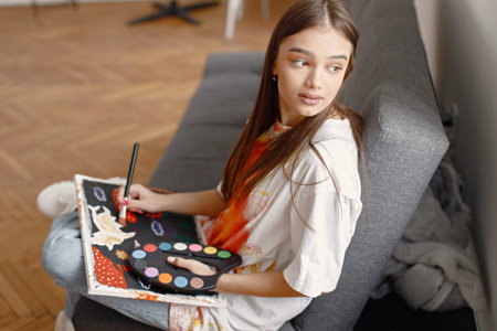 Female artist sitting on a sofa near white wall holding a canvas and brushes