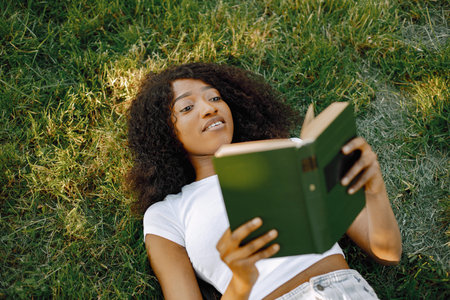 Female african student reading a book while lying on a grass in a parkの写真素材