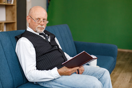 Mature man reading a book and drinking a tea on the sofa in living roomの写真素材