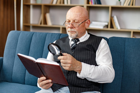 Mature man reading a book with magnifying glass on the sofa in living roomの写真素材