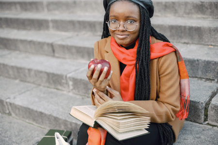 Young black woman in a coat sitting on a stairs with a book and apple