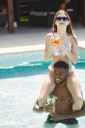Young girl and black male friend relaxing in a swimming pool and drinking a cocktails