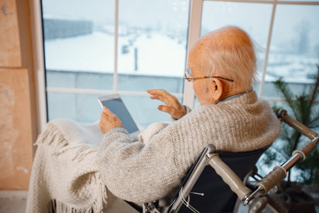Old man on a wheelchair sitting near big window and using a tabletの写真素材