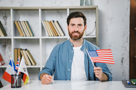 Bearded male tutor sitting at table holding flag of USA, looking at cameraの写真素材